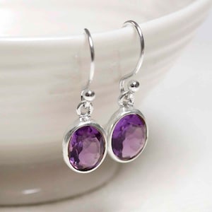 May include: Silver drop earrings featuring oval, faceted amethyst gemstones. The earrings have a simple design with a hook fastening. The purple gemstones are set in a silver bezel.