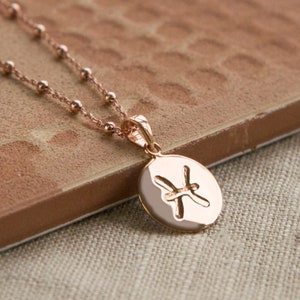 May include: Rose gold necklace featuring a pendant with the Pisces zodiac symbol. The pendant is oval-shaped and hangs from a delicate beaded chain. The necklace is displayed on a textured, brown surface.