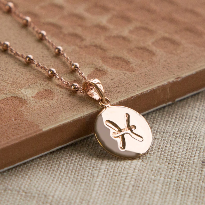May include: Rose gold necklace featuring a pendant with the Pisces zodiac symbol. The pendant is oval-shaped and hangs from a delicate beaded chain. The necklace is displayed on a textured, brown surface.