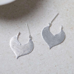 May include: A pair of silver earrings. Each earring has a heart-shaped pendant with a textured finish. The earrings have a post and a small ball detail at the top. The background is a light, neutral colour.