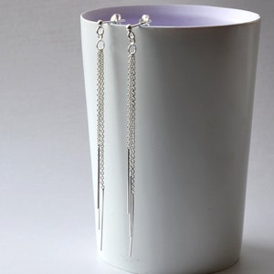 Può includere: Un paio di orecchini pendenti argentati. Ogni orecchino presenta un piccolo perno, una delicata catena e tre barre d'argento di diverse lunghezze. Gli orecchini sono esposti su sfondo bianco.