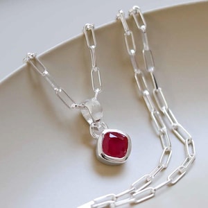 Op de afbeelding: Een zilveren ketting met een vierkante, robijnrode hanger. De hanger is gezet in een zilveren bezel en hangt aan een zilveren ketting met langwerpige schakels. De ketting wordt getoond op een lichtgekleurd oppervlak.