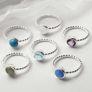 Könnte beinhalten: Eine Auswahl an Silberringen mit verschiedenen Edelsteinen, darunter Türkis, Mondstein, Amethyst und Opal. Jeder Ring hat ein Perlenband und eine runde Fassung. Die Ringe werden auf einer weißen Oberfläche präsentiert.