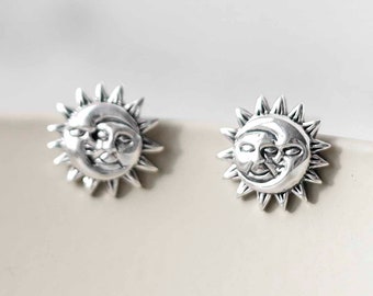 May include: A pair of silver-toned stud earrings. Each earring features a smiling sun and moon design. The sun has pointed rays around the face, and the moon is nestled within the sun's face. The earrings are set against a neutral background.