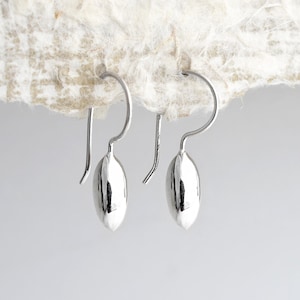 May include: A pair of silver-colored earrings. Each earring features a teardrop-shaped charm suspended from a curved hook. The earrings are displayed against a textured, off-white background.