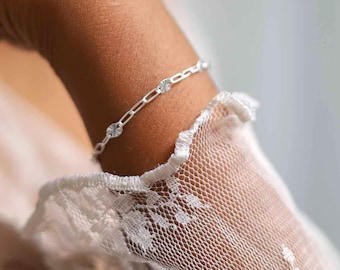 Puede incluir: Una delicada pulsera de plata con un diseño de eslabones y pequeños abalorios redondos y texturizados. La pulsera se lleva en una muñeca, con una manga de encaje blanco en primer plano.