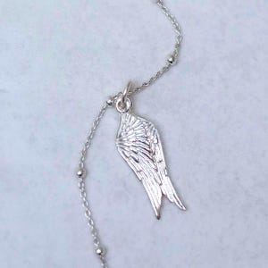 May include: A silver necklace with a wing-shaped pendant. The chain has small, spherical beads. The wing pendant is finely detailed, showing individual feather patterns. The necklace is a delicate piece of jewellery.
