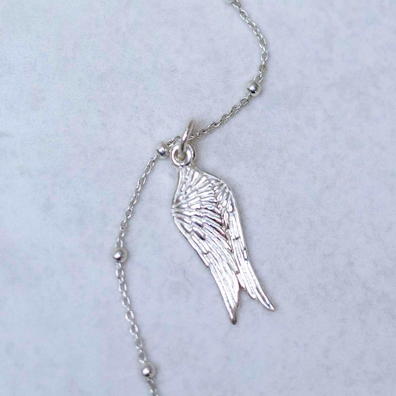 May include: A silver necklace with a wing-shaped pendant. The chain has small, spherical beads. The wing pendant is finely detailed, showing individual feather patterns. The necklace is a delicate piece of jewellery.