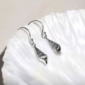 Peut inclure: Boucles d'oreilles coquillage argentées. Chaque boucle d'oreille présente un pendentif coquillage conique détaillé, suspendu à un crochet incurvé. Les boucles d'oreilles sont présentées sur une surface blanche texturée.