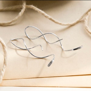 May include: A pair of silver earrings with a minimalist, abstract design. The earrings are made of thin, curved metal, with a flattened, textured end. They rest on a light brown surface, with a length of twine in the background.