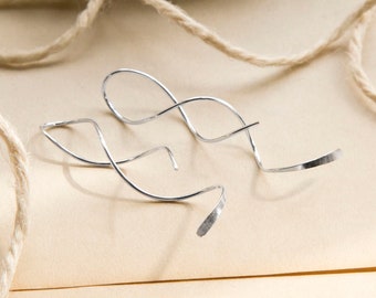 May include: A pair of silver-toned, minimalist earrings with a flowing, abstract design. The earrings are made of thin, curved metal, with a flattened, textured end. They rest on a light brown surface, with a length of twine in the background.