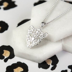 Puede incluir: Un collar plateado con un colgante con forma de cabeza de leopardo. El colgante tiene un diseño calado, que muestra los rasgos faciales del leopardo. El collar se exhibe sobre una superficie blanca con un fondo de estampado de leopardo.