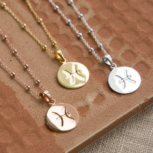 May include: Three necklaces with round pendants featuring the Pisces zodiac symbol. The necklaces are in rose gold, gold, and silver tones. Each pendant is attached to a delicate chain with small beaded accents. The necklaces rest on a textured, brown surface.