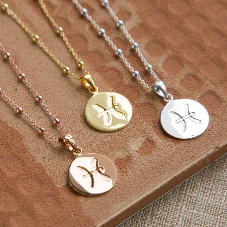 May include: Three necklaces with round pendants featuring the Pisces zodiac symbol. The necklaces are in rose gold, gold, and silver tones. Each pendant is attached to a delicate chain with small beaded accents. The necklaces rest on a textured, brown surface.