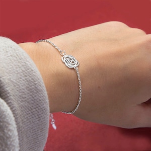 May include: A silver bracelet featuring a geometric rose design. The delicate chain bracelet is shown on a wrist against a red background. The bracelet's focal point is a stylized flower with a square center.