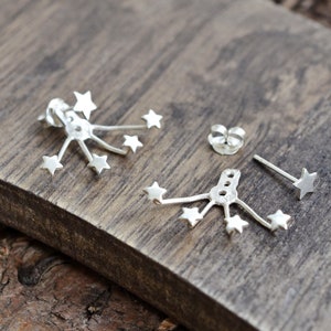 May include: Silver starburst earrings featuring multiple small stars radiating from a central point. One earring is a stud, and the other has a post with multiple stars. The earrings are displayed on a wooden surface.