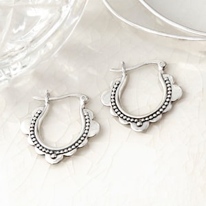 May include: A pair of silver-colored hoop earrings. Each earring features a scalloped edge design with a dotted border. The earrings are displayed on a white, textured surface.