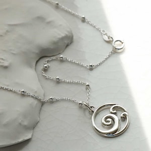 Puede incluir: Un collar de plata con un colgante con un dise&ntilde;o en espiral. El collar tiene una cadena delicada con peque&ntilde;as cuentas espaciadas uniformemente. El colgante es un marco circular con una forma estilizada de ola o espiral. El cierre es visible en la parte superior.