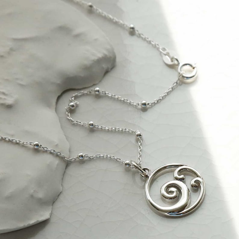 Puede incluir: Un collar de plata con un colgante con un dise&ntilde;o en espiral. El collar tiene una cadena delicada con peque&ntilde;as cuentas espaciadas uniformemente. El colgante es un marco circular con una forma estilizada de ola o espiral. El cierre es visible en la parte superior.