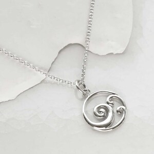Puede incluir: Un collar con colgante plateado con un dise&ntilde;o circular con un patr&oacute;n ondulado y arremolinado. El colgante cuelga de una delicada cadena. El collar se muestra sobre un fondo blanco.
