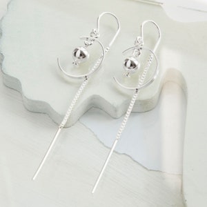 Peut inclure: Paire de boucles d'oreilles argentées au design original. Chaque boucle comprend un petit charme rond, un croissant incurvé, une chaîne délicate et une barre droite. Présentées sur une surface blanche.