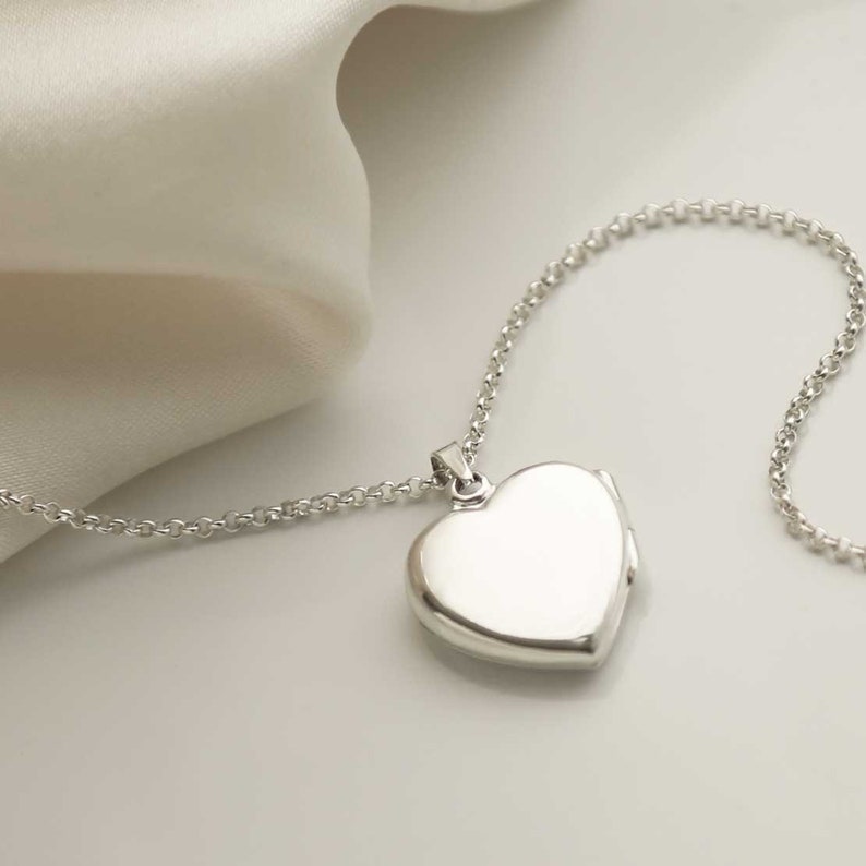 May include: A silver heart-shaped locket necklace. The locket is smooth and reflective, suspended from a delicate chain. The necklace is displayed on a white surface with a soft, draped fabric in the background.