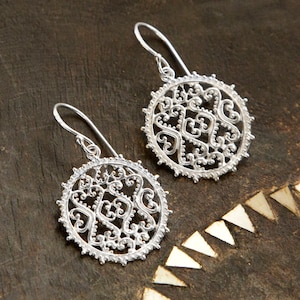 May include: A pair of silver filigree earrings with a circular design. The earrings have a delicate, intricate pattern of swirls and hearts.