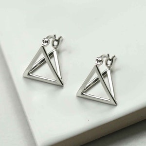 May include: A pair of silver-coloured geometric triangle hoop earrings. The earrings have a triangular design with a hinged closure. The earrings are displayed on a white surface.