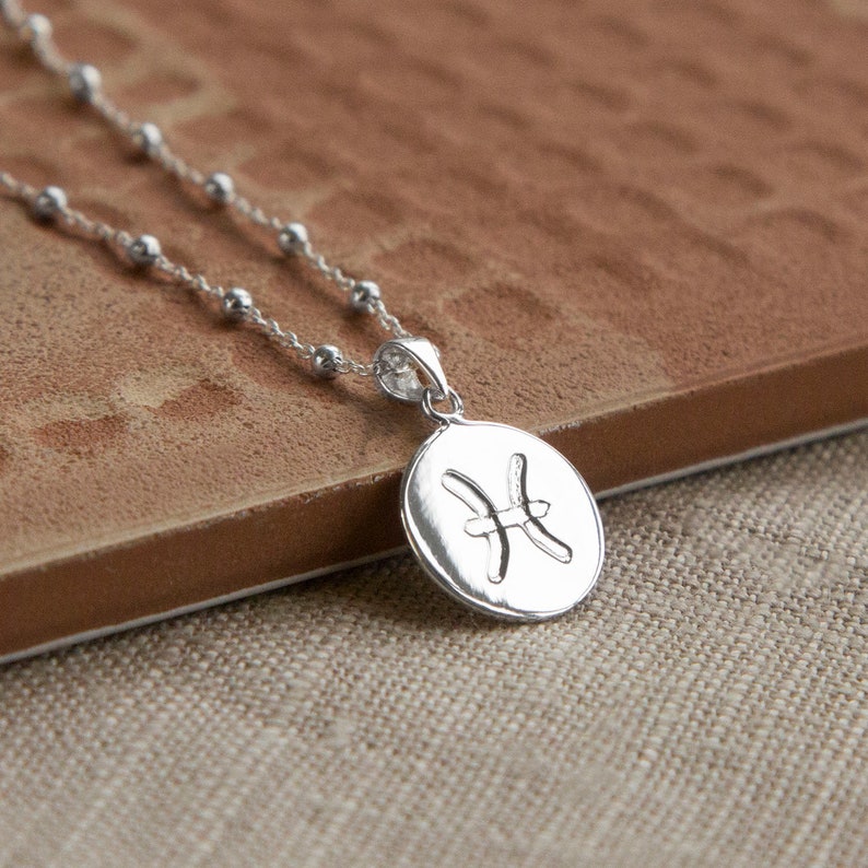 May include: A silver-colored necklace featuring a round pendant with the Pisces zodiac symbol. The pendant hangs from a delicate chain with small, evenly spaced beads. The necklace is displayed on a brown surface.