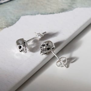 Puede incluir: Dos pendientes de botón plateados con forma de calavera. Cada pendiente presenta un diseño detallado de calavera con dientes y cuencas de los ojos visibles. Los pendientes se muestran sobre una superficie blanca, mostrando su intrincada artesanía.