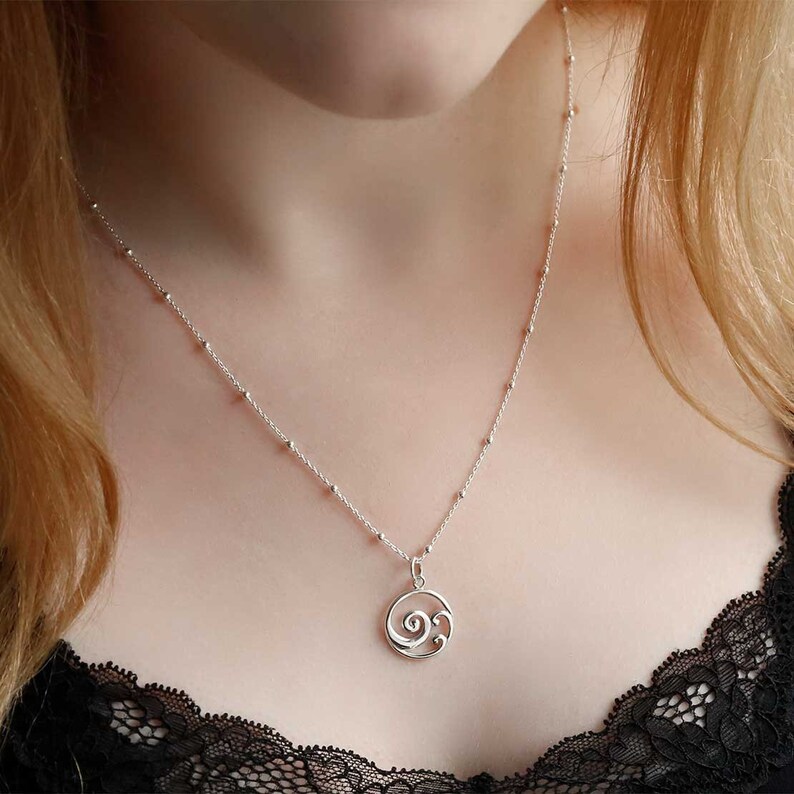 Puede incluir: Un collar de plata con una cadena delicada y un colgante con un dise&ntilde;o en espiral. El collar tiene peque&ntilde;as cuentas espaciadas a lo largo de la cadena. El colgante es una forma estilizada de ola o espiral.