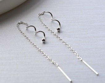 May include: A pair of silver threader earrings. Each earring features a curved design with a small ball and a long, thin bar. The earrings are displayed on a light-colored surface.