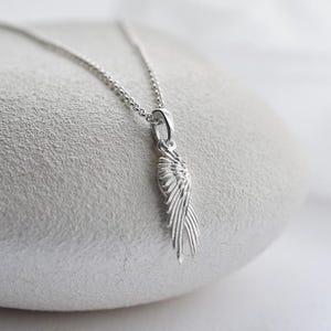 May include: A silver wing pendant necklace. The pendant, shaped like a detailed wing, hangs from a fine chain. The necklace is presented on a light, textured surface, highlighting the silver's shine. The necklace is a delicate piece of jewellery.