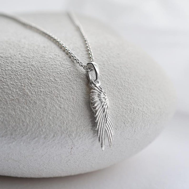 May include: A silver wing pendant necklace. The pendant, shaped like a detailed wing, hangs from a fine chain. The necklace is presented on a light, textured surface, highlighting the silver's shine. The necklace is a delicate piece of jewellery.