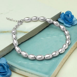 Puede incluir: Un brazalete plateado con cuentas ovaladas. El brazalete está hecho de un metal brillante y tiene un cierre de cadena ajustable. Las cuentas están espaciadas uniformemente, creando un aspecto liso. El brazalete se exhibe sobre una superficie turquesa.