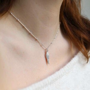 May include: A silver necklace with a detailed wing pendant. The chain is a fine silver with small beads. The wing pendant is intricately designed, showing individual feather details. The necklace is worn against a white sweater. The chain is measured in centimetres.