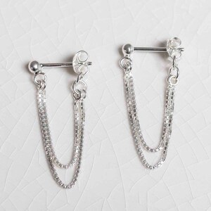 May include: A pair of silver-colored chain earrings. Each earring features a post with a small ball at the end, a butterfly backing, and two delicate, dangling chains. The chains are made of small, square links.