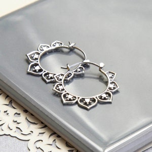 May include: A pair of silver-colored hoop earrings. Each earring features a floral design with heart-shaped petals and small circular accents. The earrings are displayed on a reflective, gray surface.