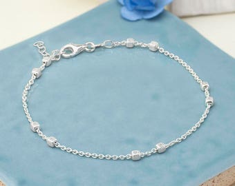 Puede incluir: Una delicada pulsera de plata con una cadena de pequeños eslabones redondos y cuentas cúbicas texturizadas espaciadas uniformemente. La pulsera tiene un cierre de mosquetón y una longitud ajustable. La joya se exhibe sobre una superficie azul claro.