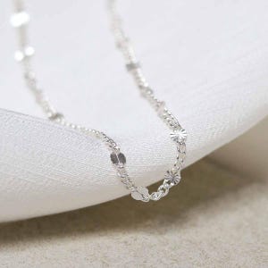 May include: A delicate silver necklace with a chain featuring small, round, and flower-shaped charms. The necklace is displayed on a white, textured surface, highlighting its intricate design and reflective qualities. The jewelry is suitable for everyday wear.