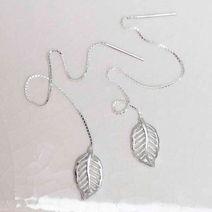 May include: Silver leaf threader earrings. These earrings feature a delicate leaf design suspended from a fine chain. The leaves have intricate detailing, and the earrings are shown against a white background.