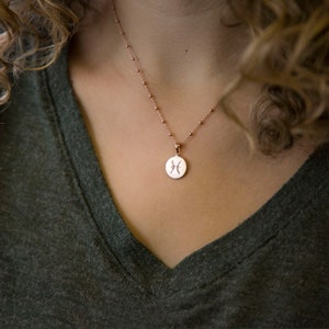 May include: Rose gold necklace featuring a round pendant with the Pisces zodiac symbol. The pendant hangs from a delicate beaded chain. The necklace is displayed against a dark green v-neck shirt.