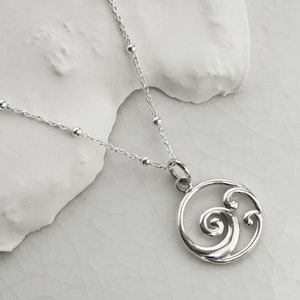 Puede incluir: Un collar de plata con un colgante de ola. El colgante es un dise&ntilde;o circular con un patr&oacute;n de ola estilizado. La cadena es de plata delicada con peque&ntilde;os detalles de cuentas. El collar se muestra sobre una superficie blanca.