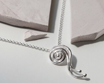 Puede incluir: Un collar con colgante plateado con un diseño en espiral y dos extensiones curvas que terminan en pequeñas esferas. El colgante cuelga de una delicada cadena. El collar se muestra sobre un fondo neutro con una piedra rota.