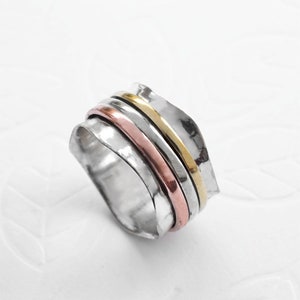 May include: A silver-toned ring with a textured band and three thinner bands in gold, rose gold, and silver. The ring has a unique, layered design, and the bands appear to spin freely. The ring is set against a white background.