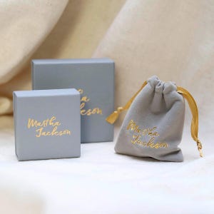 Op de afbeelding: Drie sieradenverpakkingen: twee grijze dozen en een grijze trekkoordtas. De dozen en de tas hebben gouden tekst met de tekst "Martha Jackson". De tas heeft een gouden trekkoord. De achtergrond is een zachte, gebroken witte stof.