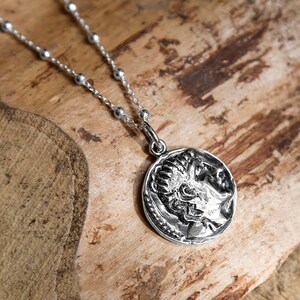 Puede incluir: Un collar de cadena de plata con un colgante de moneda de plata que presenta un perfil de un rostro. La moneda está detallada con un borde elevado y diseños intrincados.