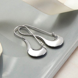 May include: A pair of polished silver earrings. The earrings have a teardrop shape with a curved, open design. The earrings are displayed on a grey surface with a white ribbon in the background. Jewellery.