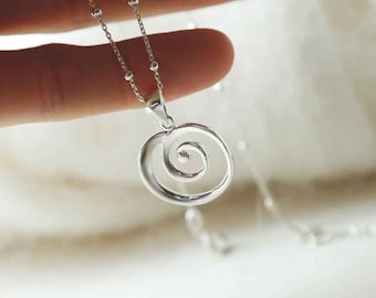 May include: A silver pendant necklace featuring a spiral design. The pendant hangs from a delicate chain with small, round beads. The necklace is shown against a soft, blurred background, highlighting the jewelry's intricate details.