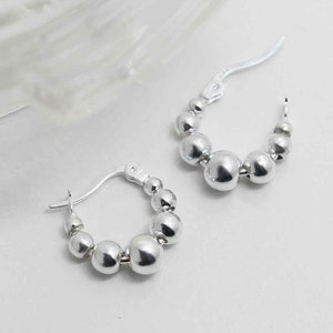 May include: A pair of silver hoop earrings with a row of round beads. The earrings are made of metal and have a polished finish.